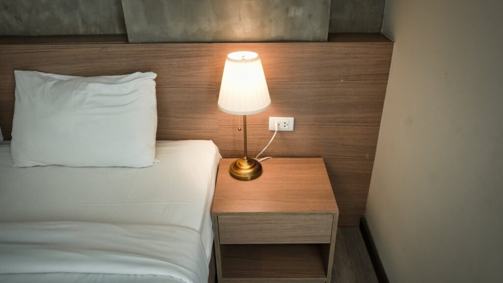 A bedside lamp illuminating a wooden nightstand beside white bedding