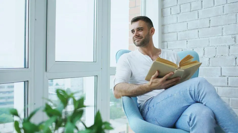 Man holding an open book while seated beside a window