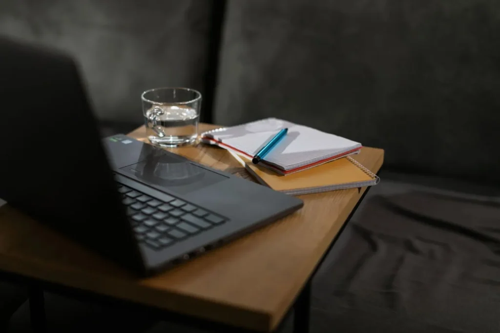 Notebook and glass of water beside a laptop on a wooden table